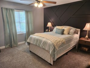 Une chambre à coucher avec une tête de lit capitonnée, un lit avec une literie à rayures, deux lampes de chevet, un ventilateur de plafond et une fenêtre avec des rideaux.