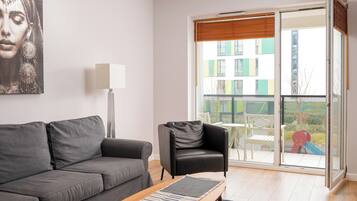 A modern living room with a grey sofa, a black leather chair, a floor lamp, and a view of a cityscape through a large sliding glass door.