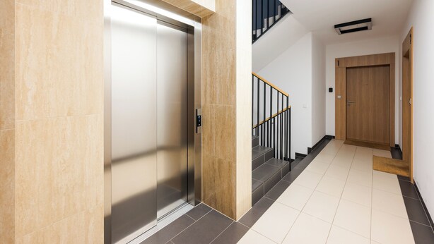 A modern elevator with a metallic finish, a wooden door frame, and a staircase with black railings leading to a second floor.