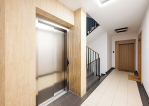 Un ascenseur moderne aux finitions métalliques, doté d’un cadre de porte en bois et d’un escalier avec des rampes noires menant à un deuxième étage.