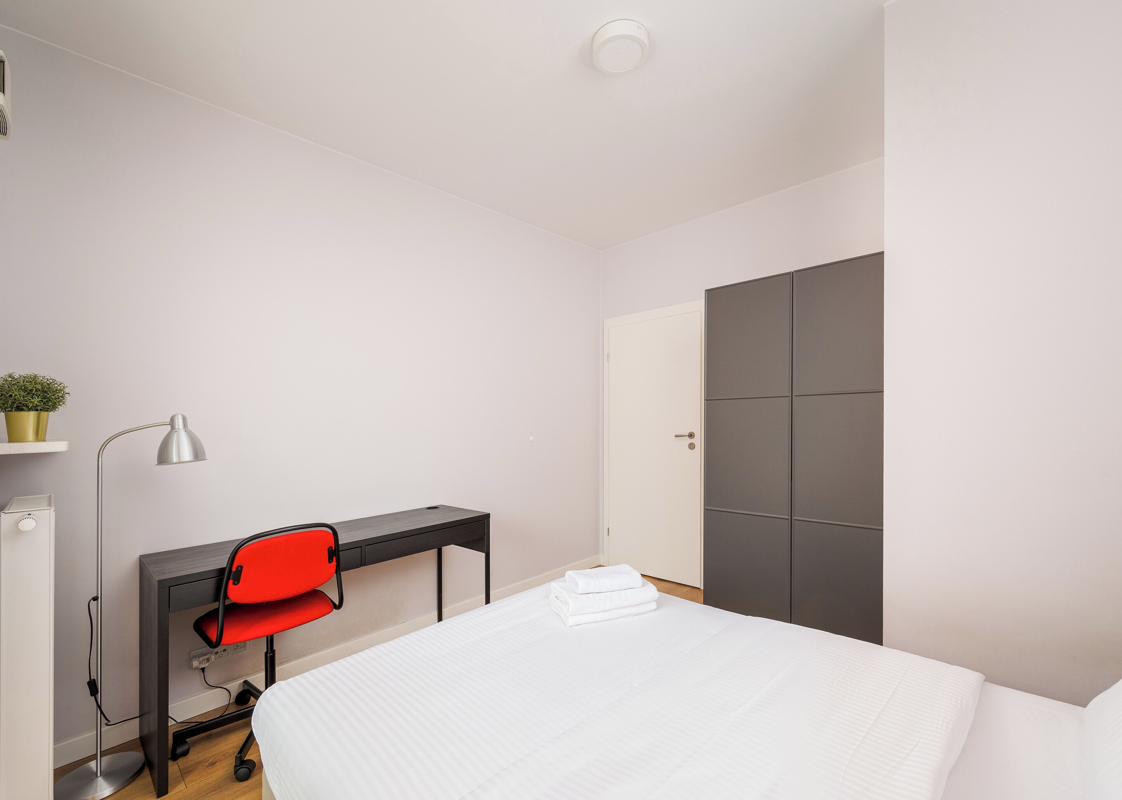 A hotel room with a bed, a desk with a chair, a lamp, and a wardrobe.