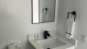 Ein Badezimmer mit einem weißen Waschbecken, einem schwarzen Wasserhahn und einem Spiegel darüber. Eine Toilette befindet sich links und ein Handtuchhalter über dem Waschbecken.