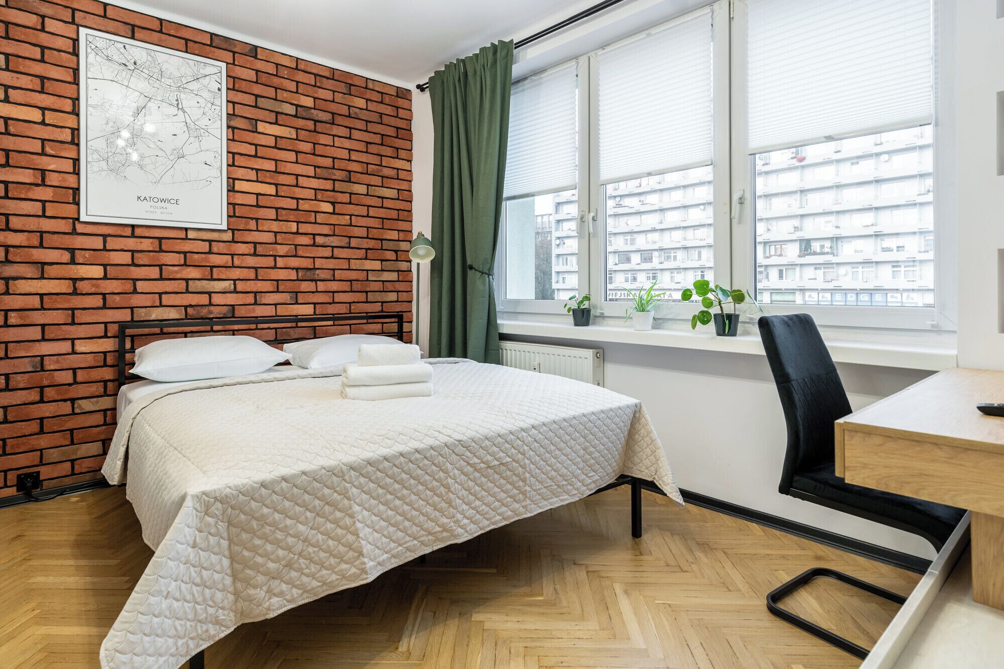 A hotel room with a brick wall, a bed with white bedding, a desk with a chair, and a window with a view of buildings.
