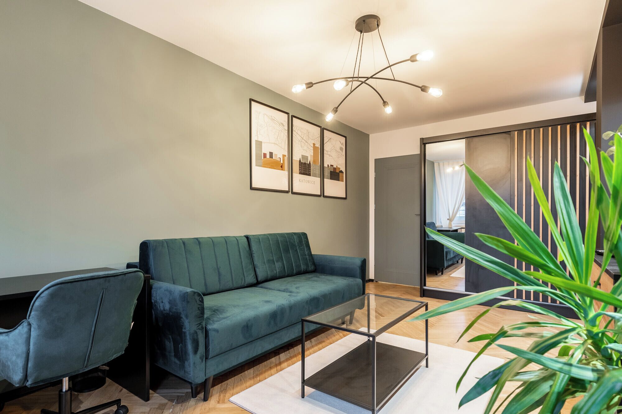 Un salon moderne avec un canapé de couleur turquoise, une table basse en verre et un bureau avec une chaise. Des tableaux représentant des paysages urbains encadrés ornent le mur, et un grand miroir reflète l’ensemble de la pièce.