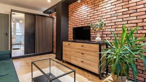 Un salon moderne avec un mur en briques, un téléviseur à écran plat, un meuble en bois, une table basse en verre et une plante en pot.