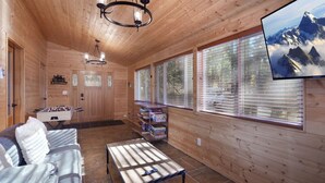 Interior de una cabaña de madera con un sofá, una mesa de centro, una mesa de futbolín y un televisor que muestra una vista de montaña.