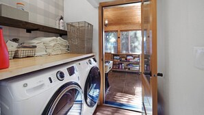 Un área de lavandería con lavadora y secadora, estantes con toallas dobladas y una puerta de madera que da a un cuarto con piso de baldosas y estanterías.