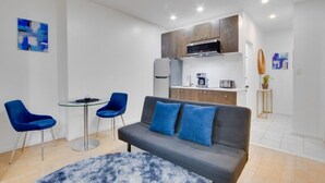 Una sala de estar moderna con un sofá gris, dos sillas azules y una mesa de centro de vidrio. Se ve una cocina al fondo con armarios de madera y un horno empotrado.