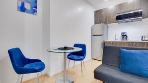 Cocina moderna con horno empotrado, microondas, cafetera y refrigerador. Cuenta con una mesa redonda de vidrio, dos sillas azules y un sofá gris con una almohada azul.