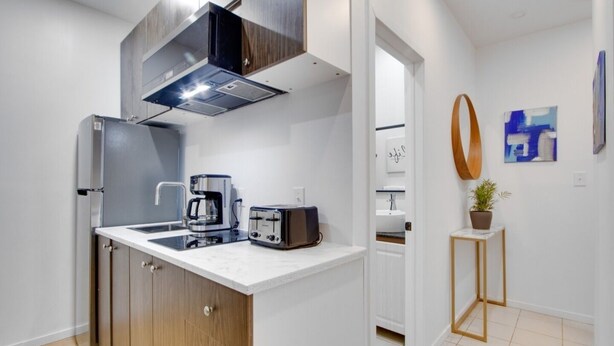Una cocina compacta con estufa integrada, cafetera y tostadora. Se observa un pequeño armario de madera y una encimera blanca.