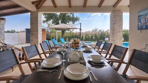 Un coin repas avec une table dressée pour six personnes, entourée de chaises, et offrant une vue sur une piscine et une végétation luxuriante en arrière-plan.