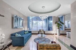 A modern living room with a blue sofa, a glass coffee table, and a large window offering a view of the outside.