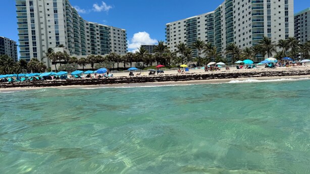 A beachfront hotel with multiple high-rise buildings, clear turquoise water, and a sandy beach with umbrellas.