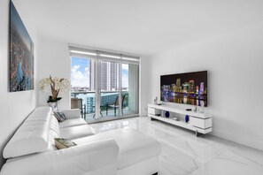 A modern living room with a white sofa, a flat-screen TV, and a cityscape view through large windows.