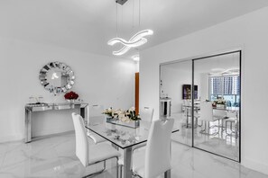 Un comedor moderno con una mesa de vidrio, sillas blancas, un espejo decorativo y vista panorámica de la ciudad a través de un panel de vidrio.