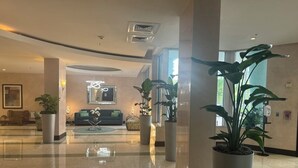 A spacious lobby with a polished floor, large potted plants, a seating area with a sofa and chairs, and a reception desk.