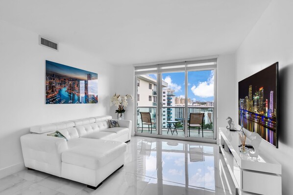 Una sala de estar moderna con un sofá blanco, un televisor de pantalla plana y un ventanal con vista a la ciudad.