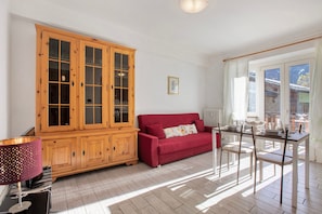 A room with a wooden cabinet, a red sofa, a dining table with chairs, and a window with curtains.