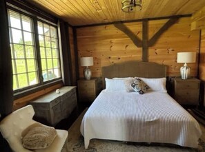 Un dormitorio con paneles de madera, una cama grande, dos mesitas de noche con lámparas, una silla y una ventana con vistas a la vegetación.