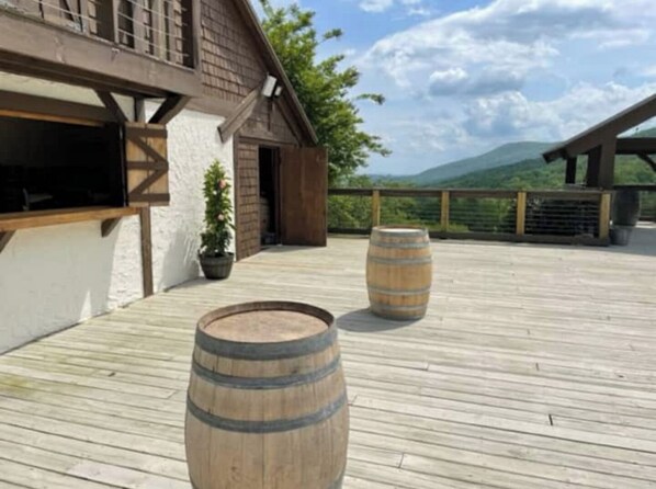 Una terraza de madera con dos barriles, un edificio de fachada blanca y un paisaje montañoso al fondo.