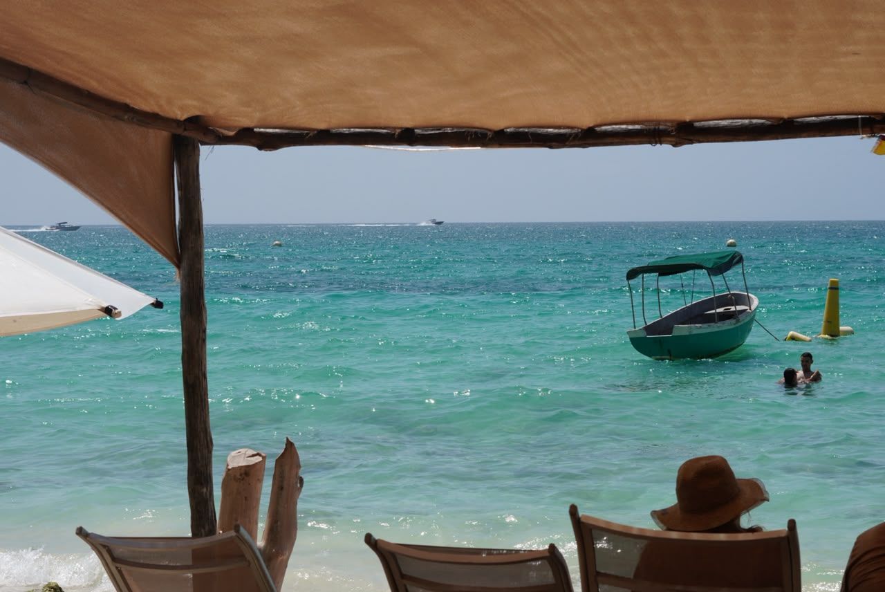 Escena de playa con un bote, una persona nadando y un área sombreada con sillas.