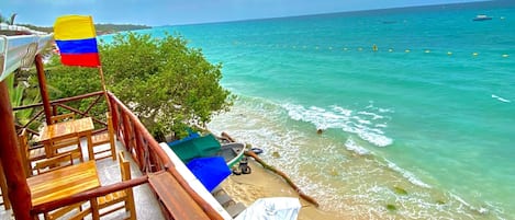 Una terraza de madera con sillas y mesas da vista a una playa con agua turquesa cristalina y una bandera.