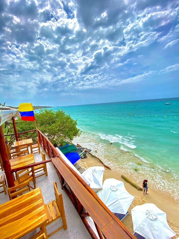 A wooden deck with chairs and tables overlooking a beach with clear turquoise water and a flag.