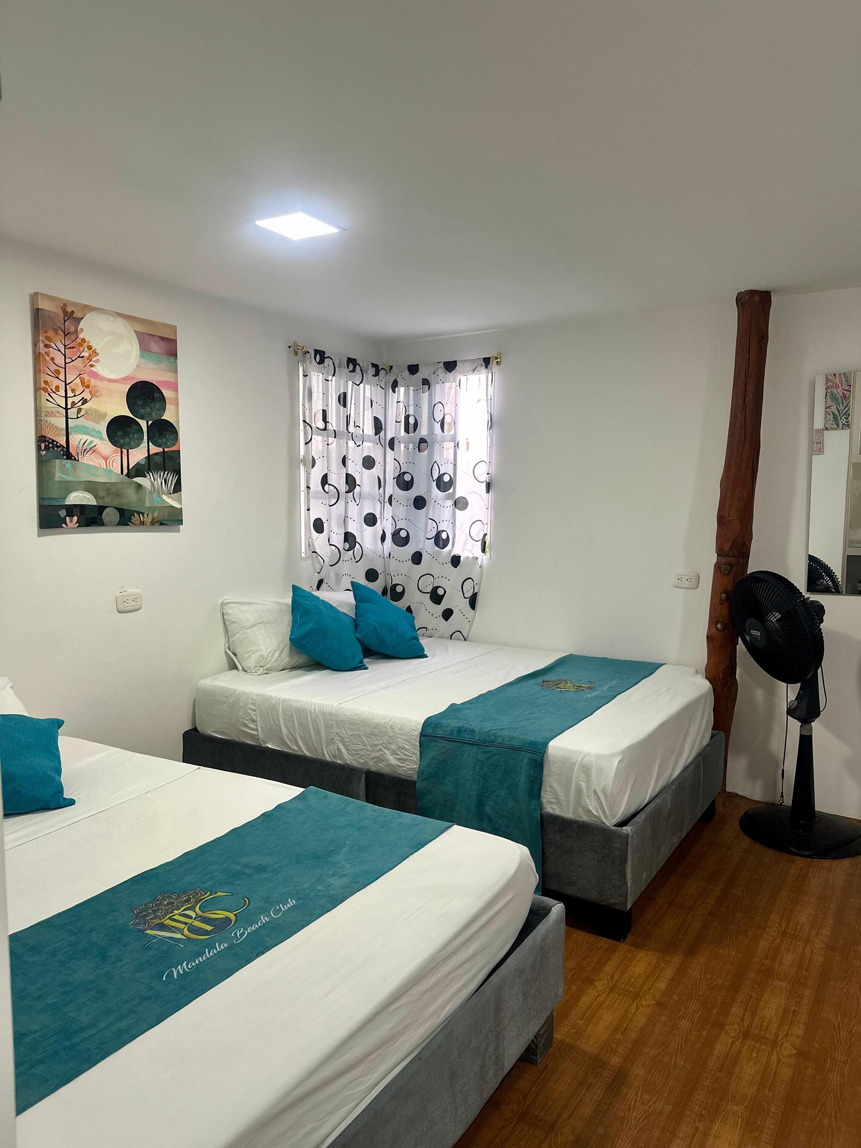 Hotelkamer met twee bedden, een plafondventilator, een schilderij aan de muur en een raam met geruit gordijnen.
