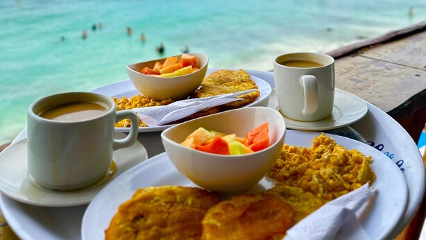 Et bord med morgenmadsprodukter, herunder æg, frugt og kaffe, med udsigt til en strand med krystalklart turkis vand og badende mennesker.