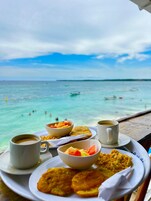 Una mesa con alimentos para el desayuno, como huevos, fruta y café, con vista a una playa de aguas turquesas cristalinas y gente nadando.