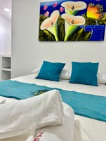 Uma cama com roupa de cama branca e duas almofadas azuis, um cobertor azul-esverdeado e uma pintura floral na parede.