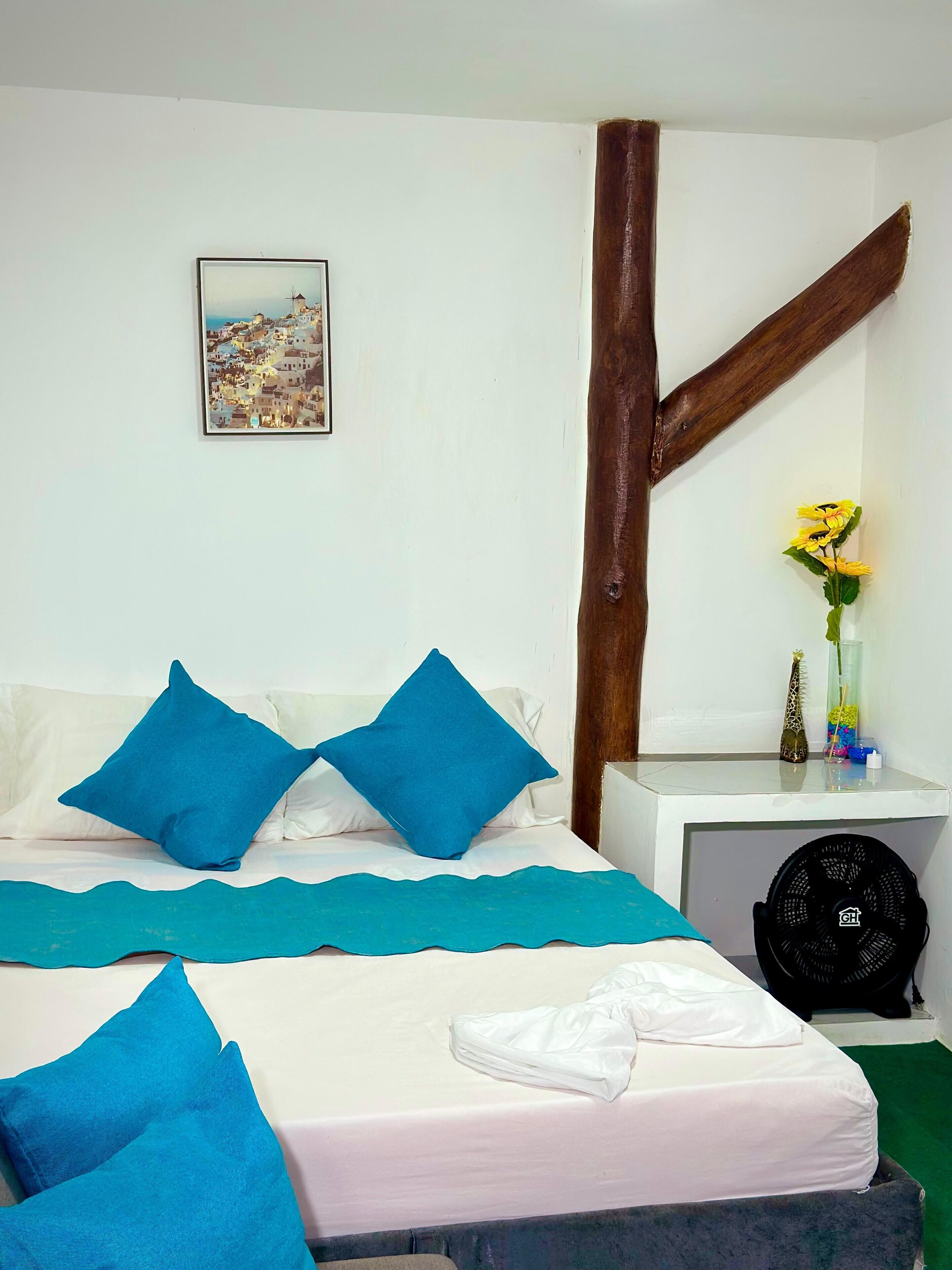 Une chambre à coucher avec un lit, des oreillers bleus, un matelas blanc, un tableau au mur, une table de chevet avec un vase et des fleurs, et un ventilateur.