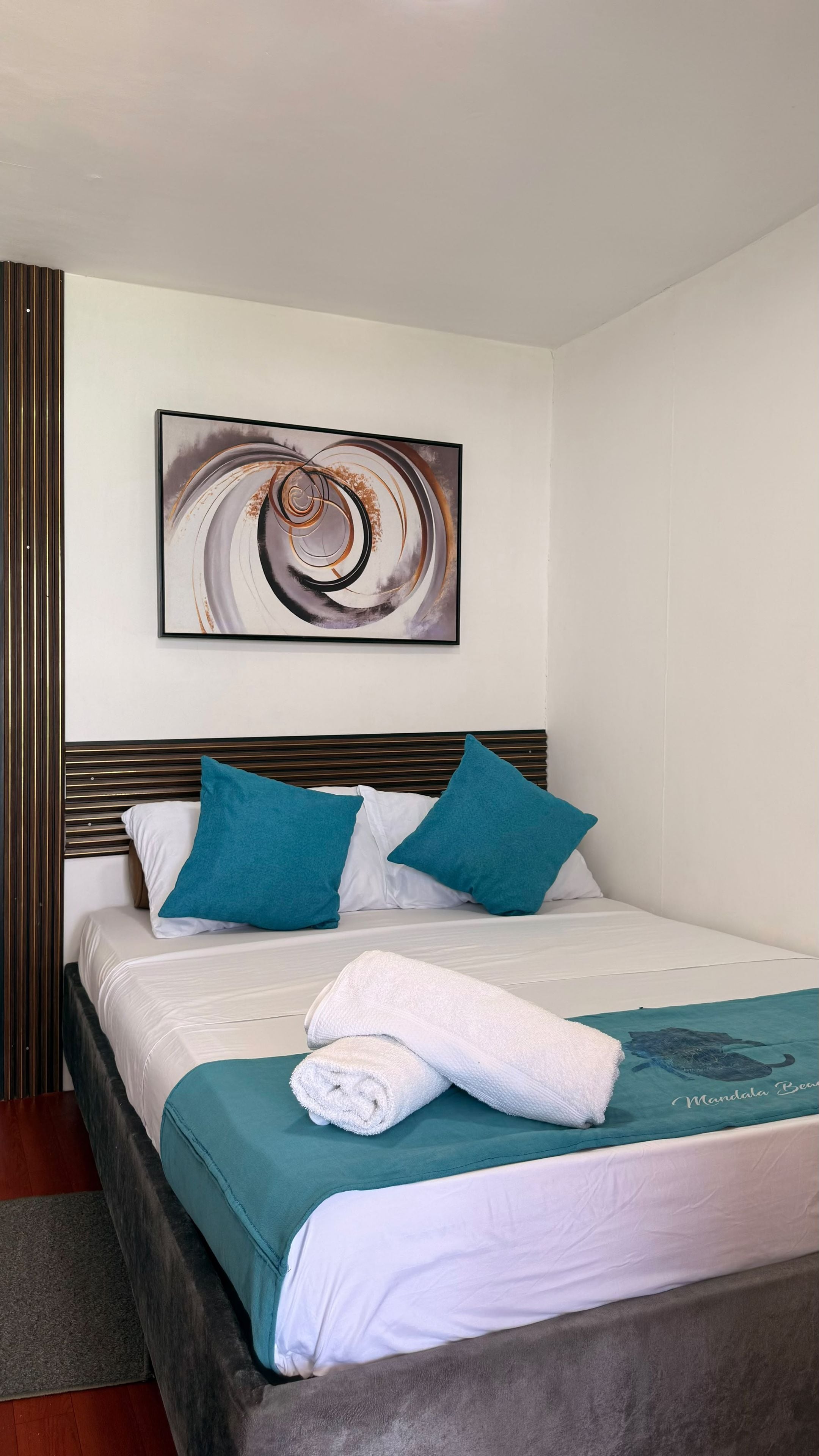 Una cama bien hecha con una colcha azul turquesa y ropa de cama blanca, una toalla blanca enrollada y dos almohadas azul turquesa.