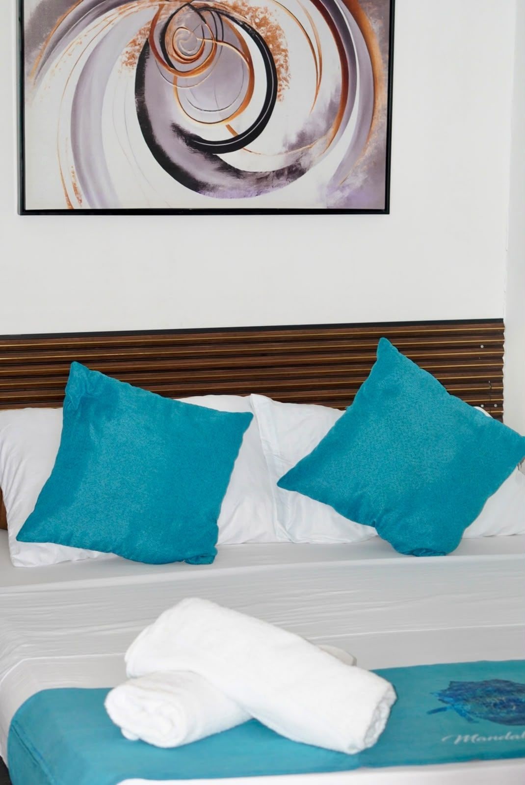 A hotel room with a bed, two teal pillows, a white blanket, and a framed abstract painting on the wall.