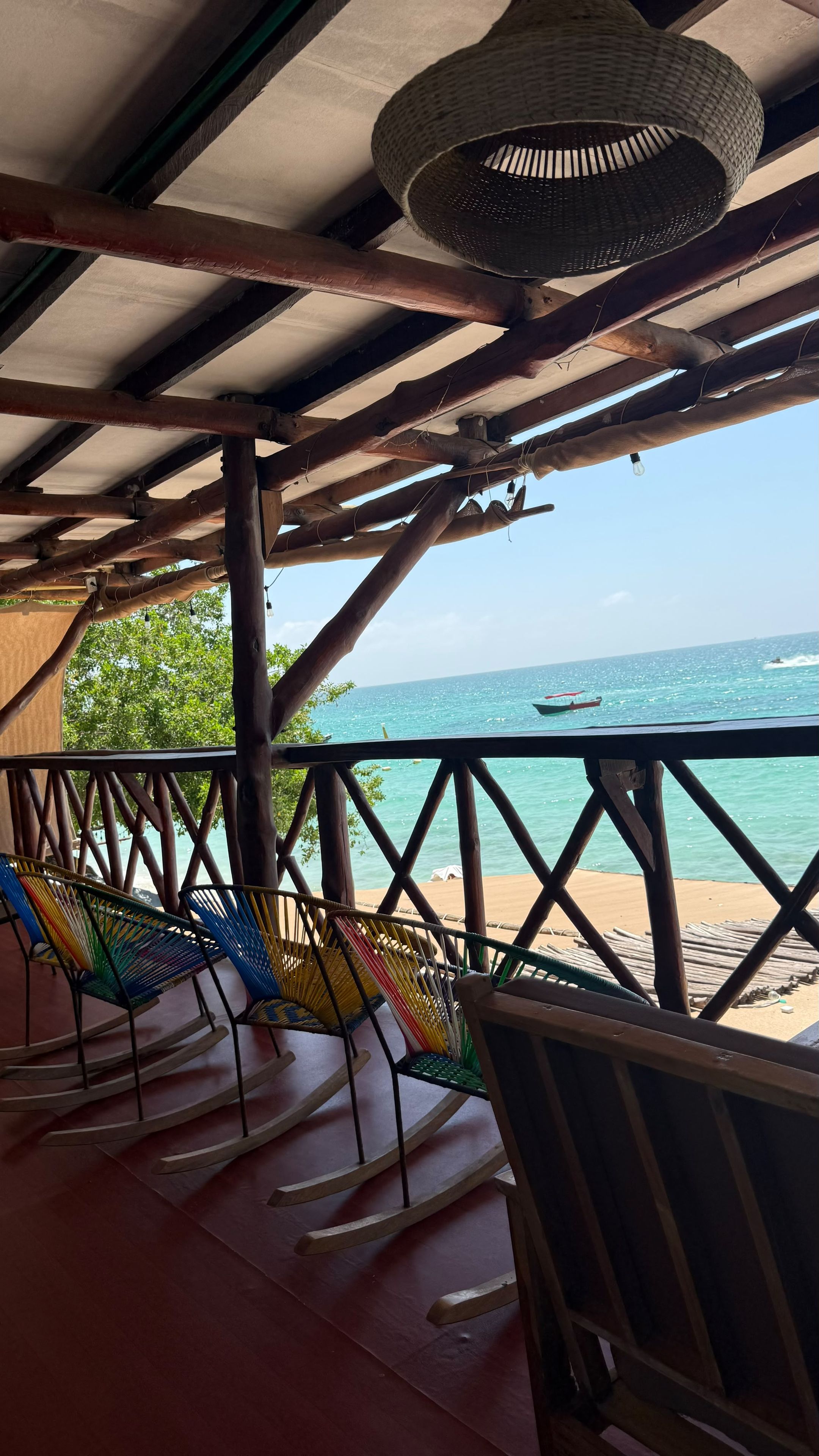 A wooden deck with a view of the sea, featuring a woven ceiling light fixture and colorful rocking chairs.