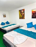 Habitación de hotel con tres camas, cada una con almohadas azules y ropa blanca. Hay dos cuadros en las paredes, uno con una vista urbana y otro un retrato.