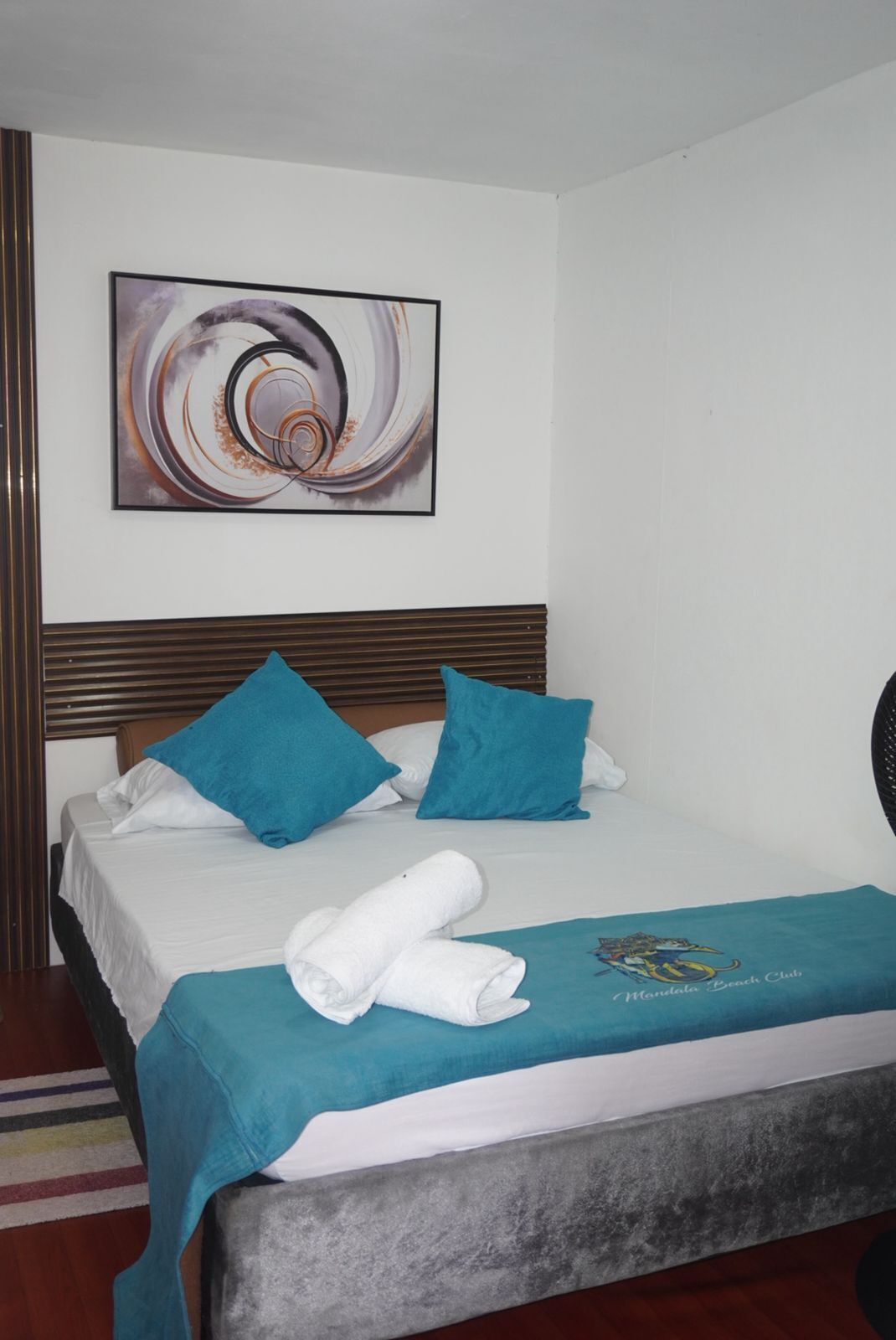 A hotel room with a bed, a headboard, a wall-mounted abstract painting, and a blue towel on the bed.