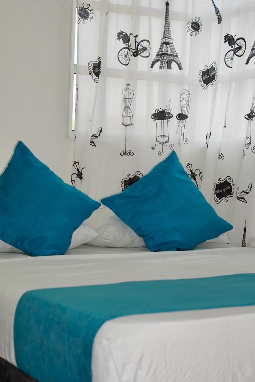 A bed with two blue pillows and a white bedspread, a window with a curtain featuring black line drawings of Parisian landmarks.