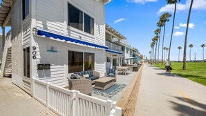 A white two-story building with a balcony, a covered patio with furniture, and a clear view of the beach and palm trees. - Beachside Oasis in Newport Beach – Steps to Balboa Park, Pier, and The Fun Zone! (Newport Beach)