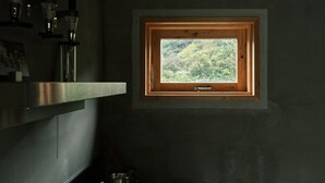 A small window with a wooden frame, a view of a forested area, and a kitchen area with a metal pot. - NEGURA (Ogimi)