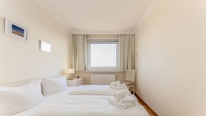 Habitación de hotel con una cama grande, una mesita de noche con lámpara, una silla y una ventana con cortinas.