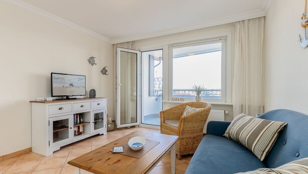 Una sala de estar con televisor, un mueble blanco, una silla de mimbre, un sofá azul y un ventanal con vistas a la ciudad.
