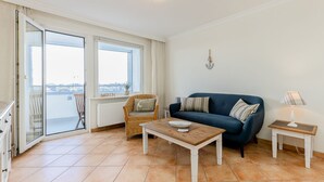 Una sala de estar con un sofá azul, una silla de mimbre y una mesa de centro de madera. Se ve el exterior a través de la puerta de vidrio.