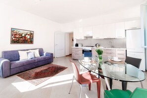 Una sala de estar moderna con un sofá, una mesa de comedor de vidrio y una zona de cocina con gabinetes y electrodomésticos blancos.