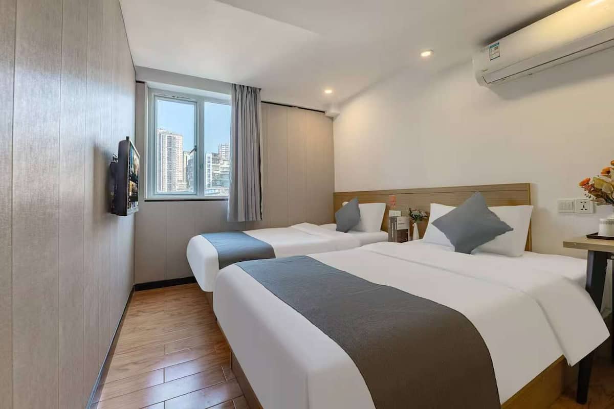A hotel room with two beds, a TV mounted on the wall, a window with curtains, and an air conditioning unit.