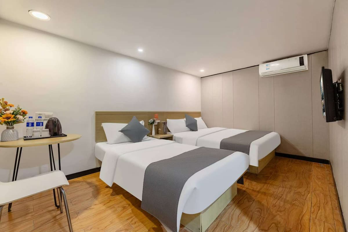 A hotel room with two beds, a small table with a vase of flowers, and an air conditioning unit on the wall.