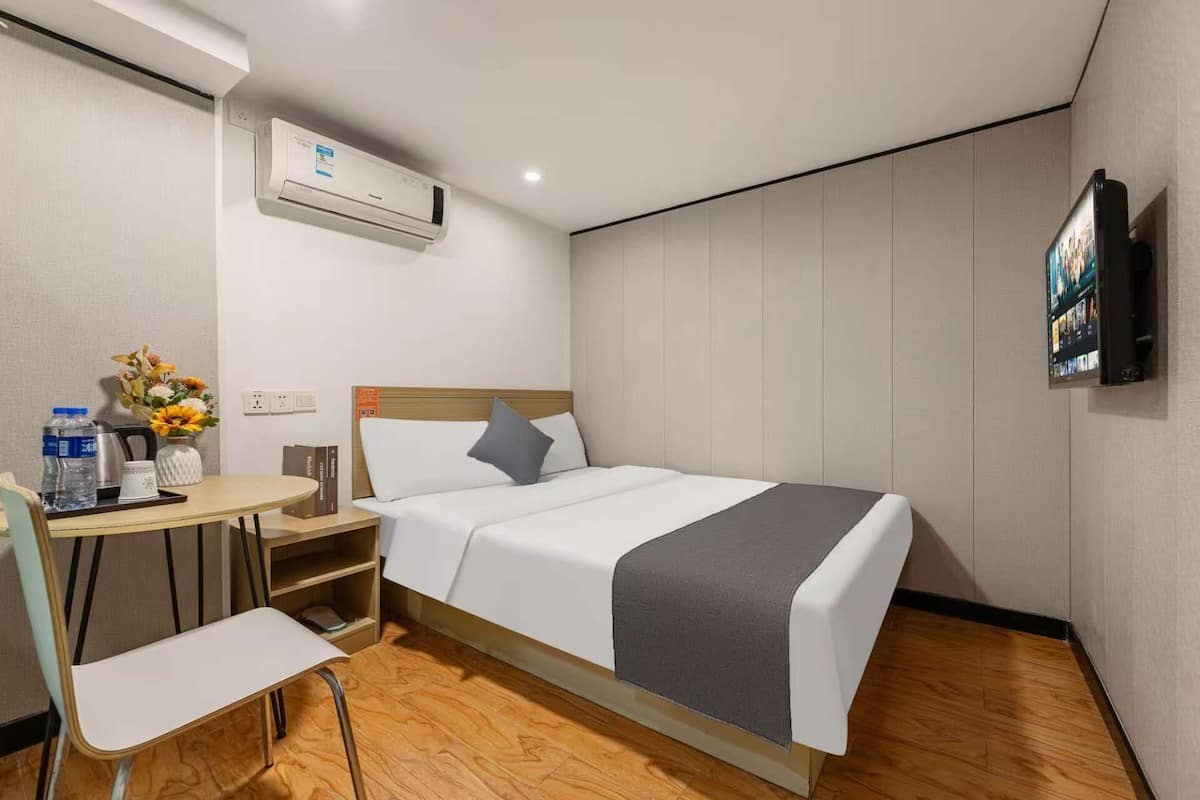A hotel room with a bed, a desk with a chair, an air conditioner, a television, and a small table with a vase of flowers.