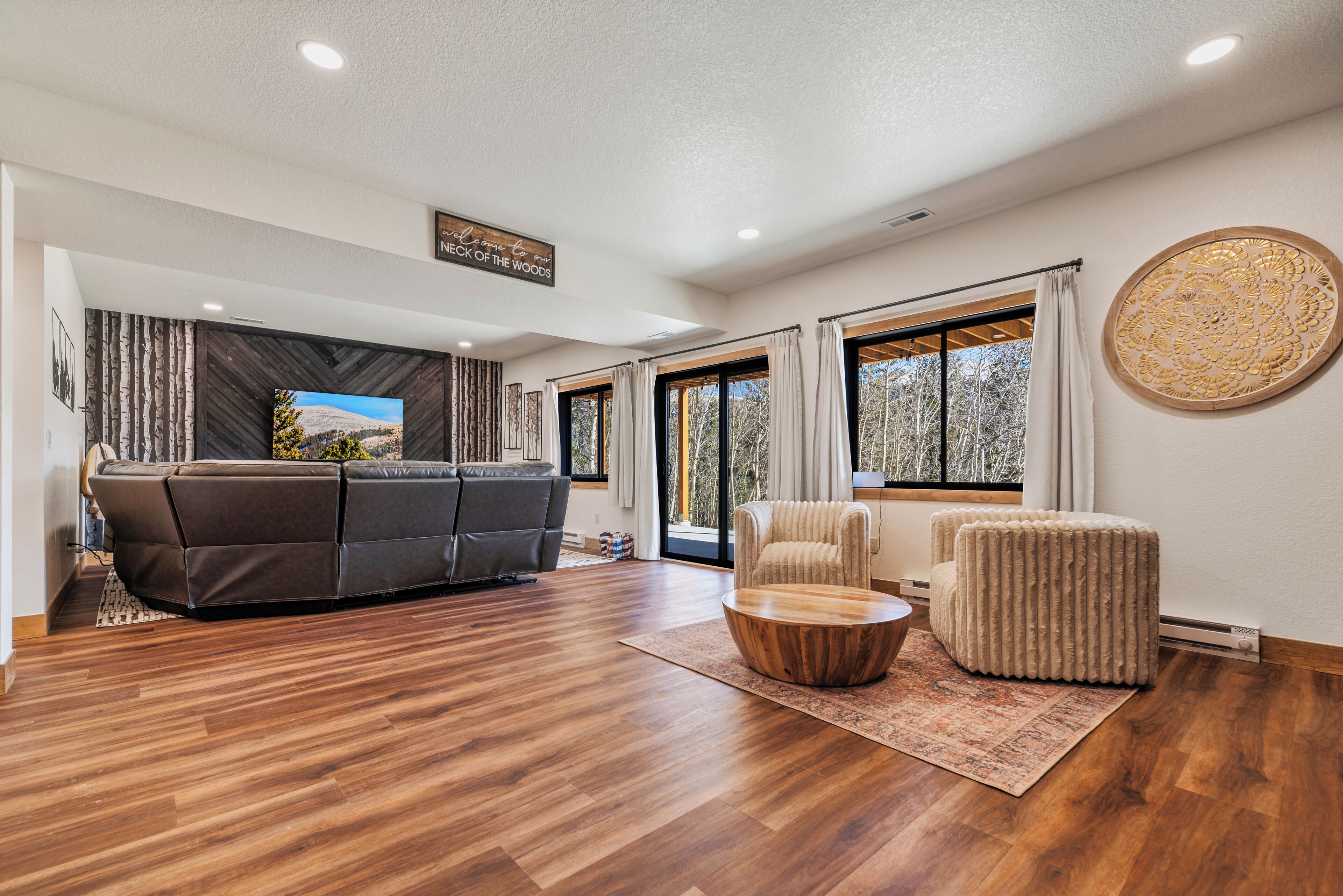 A spacious living room with a dark leather sofa, a round wooden coffee table, and a cream-colored armchair. A large window provides a view of trees outside.