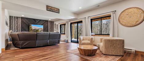 A spacious living room with a dark leather sofa, a round wooden coffee table, and a cream-colored armchair. A large window provides a view of trees outside.
