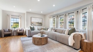A spacious living room with a large sectional sofa, a round coffee table, and a view of greenery through large windows. - Surreal Serenity Escape wIth Game Room, Pool & Spa (La Mesa)